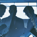 Trzech mężczyzn w garniturach rozmawia w nowoczesnym biurowcu, widoczne jako sylwetki na tle dużych okien.